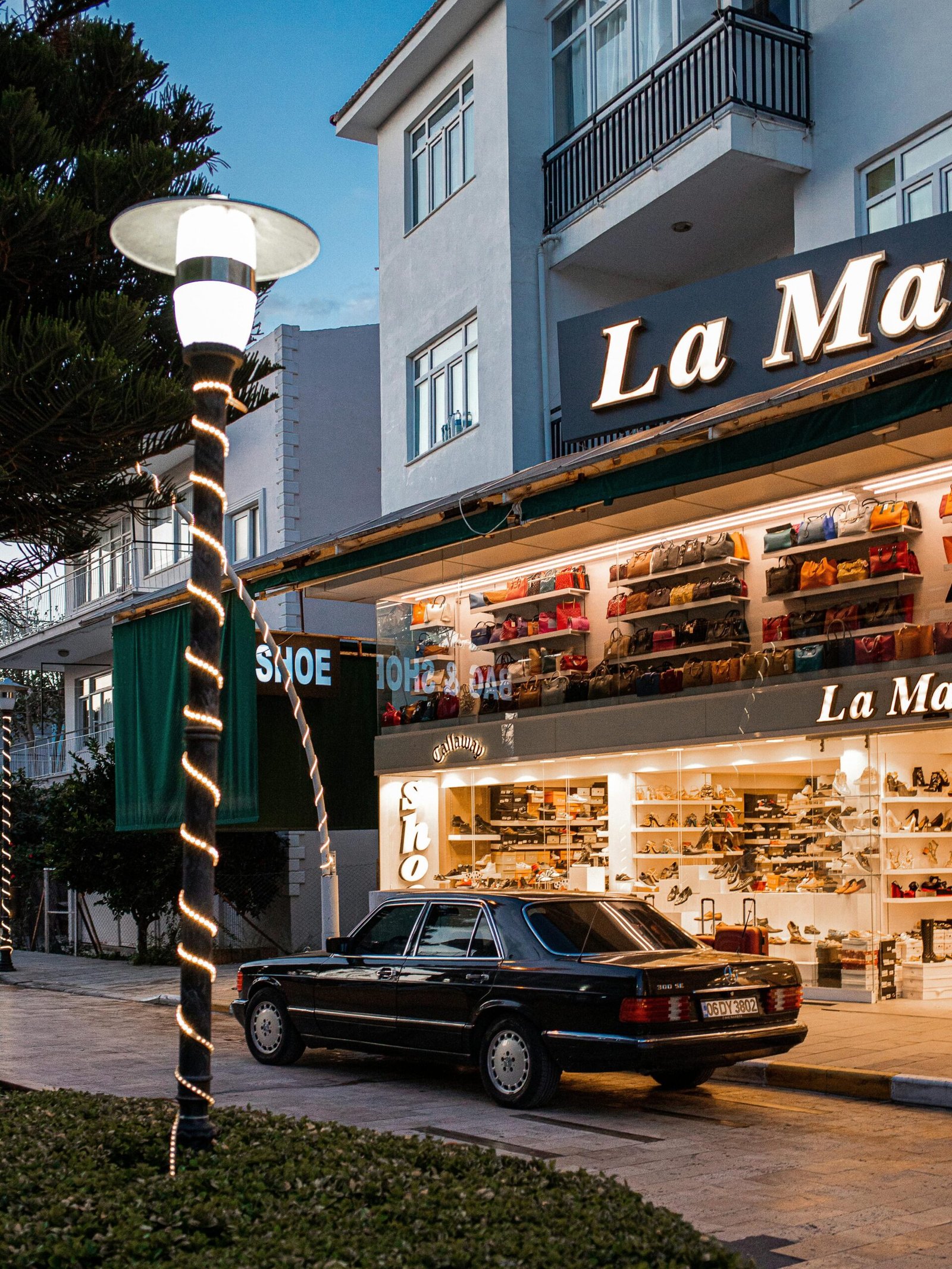 A vintage black Mercedes-Benz parked in front of a well-lit La Marca shoe store in Turkey at dusk.